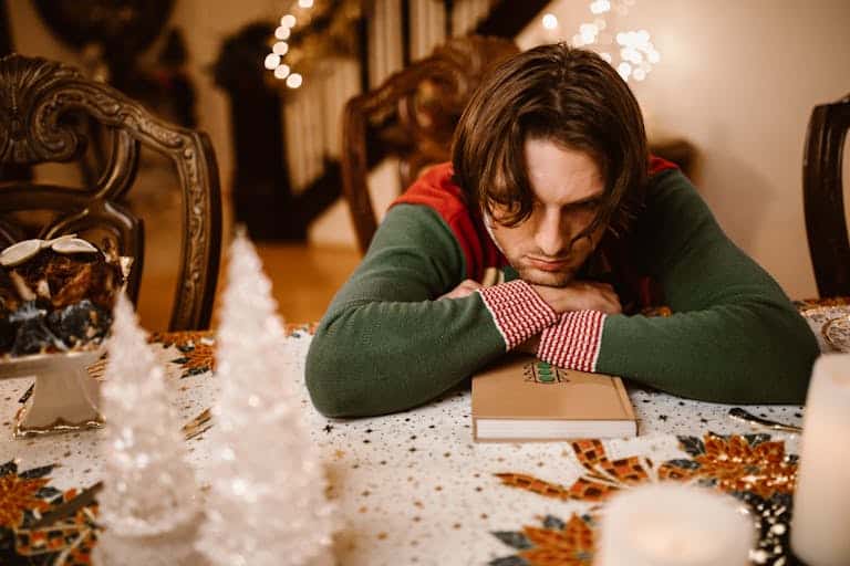 A man in a sweater looks thoughtful and sad at a decorated Christmas table indoors.